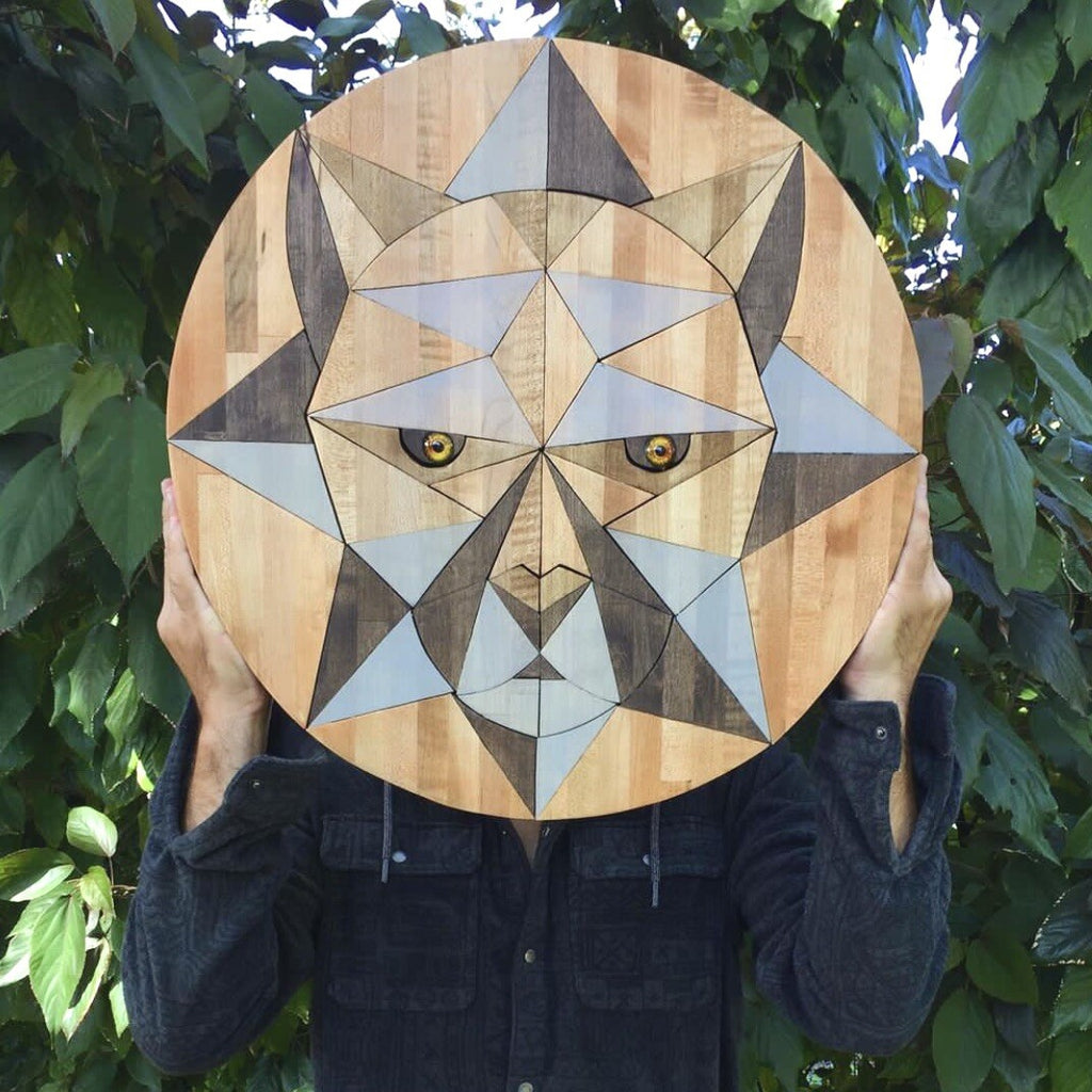 Artist person holding a wooden cutting board wood animal art with geometric mountain lion cat face design against a leafy green background